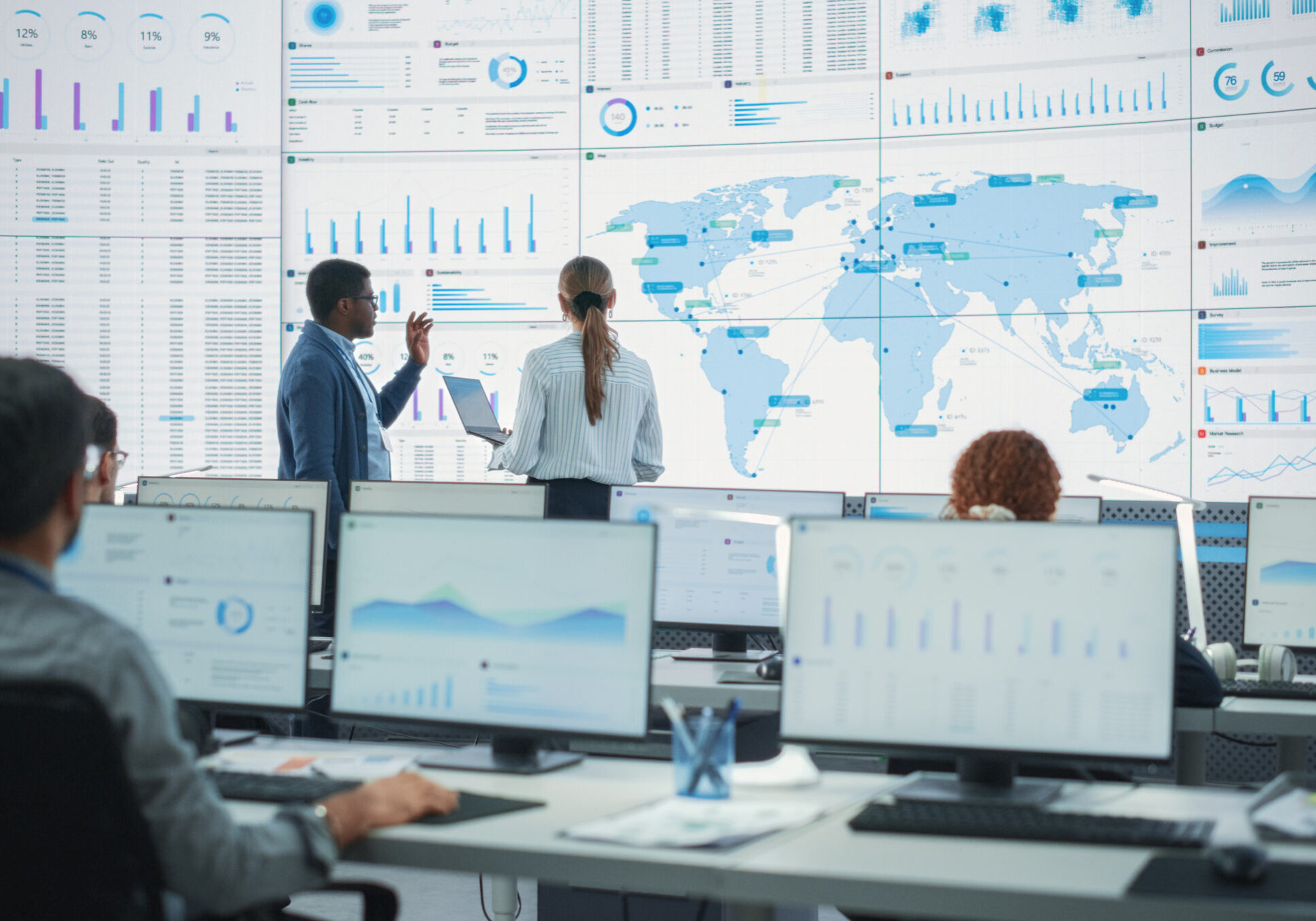 Back View Of Black Male Analyst Talking To Caucasian Female Manager In Front Of Big Digital Screen With World Map In Monitoring Office. Diverse Employees Working Behind Computers In Logistics Company.